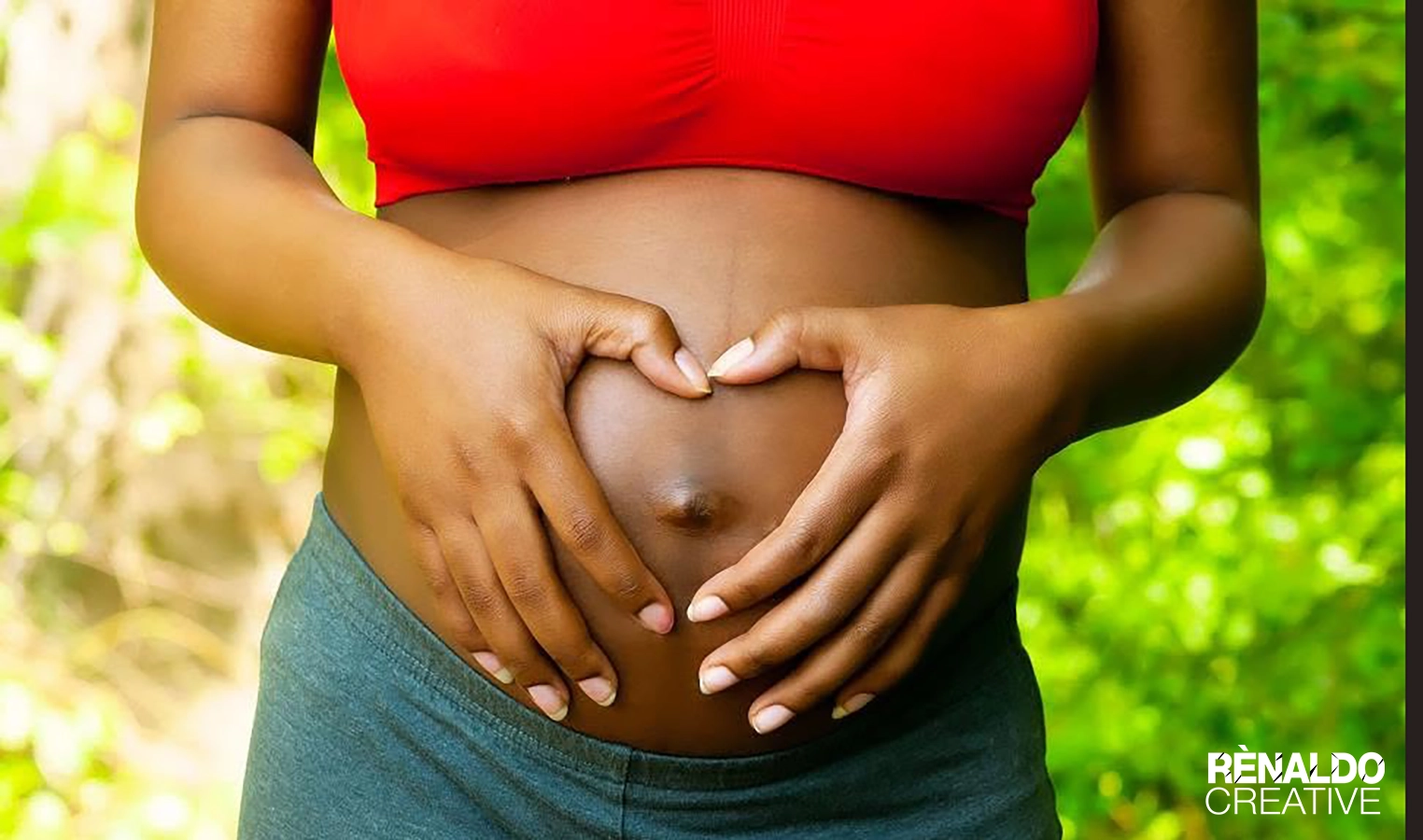 Pregnant person forming a heart shape with hands on their belly, surrounded by a natural green background.