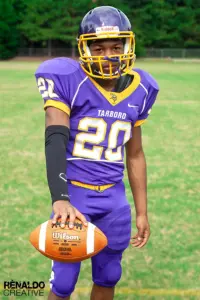 Tarboro High School varsity football player number 20 in purple and gold uniform holding a Wilson football on the field