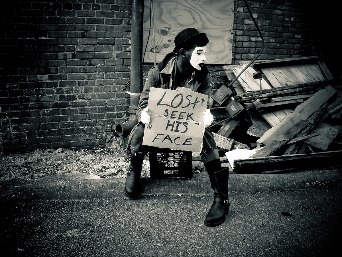 Black and white urban mime sits by a brick wall holding a cardboard sign reading "LOST? SEEK HIS FACE" amid street debris