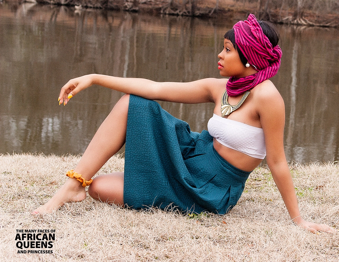 A beautiful light skin black woman in pink headwrap, green dress and white top sites on the ground next to a river looking forward Blog Header
