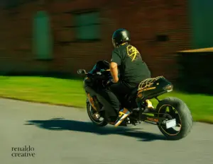 Stunt rider on black Yamaha sportbike labeled "Thelma" riding fast past brick architecture, Renaldo Creative motion portrait