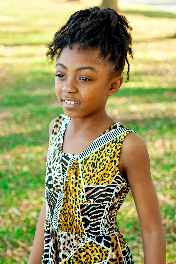 Young girl with natural hair smiles while wearing a colorful animal print dress in a sunny outdoor setting.