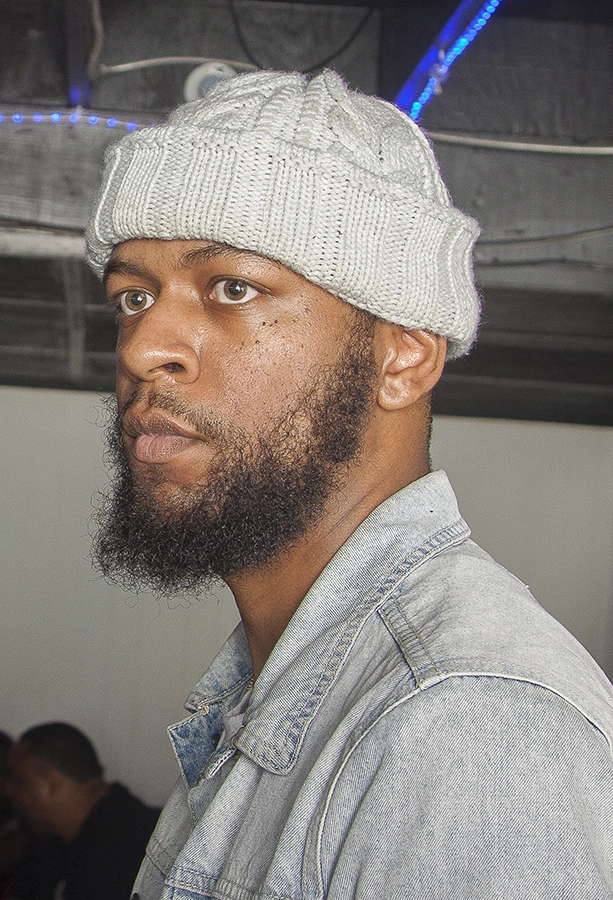 A man with a beard and gray knit hat gazes thoughtfully, wearing a denim jacket in a dimly lit setting.