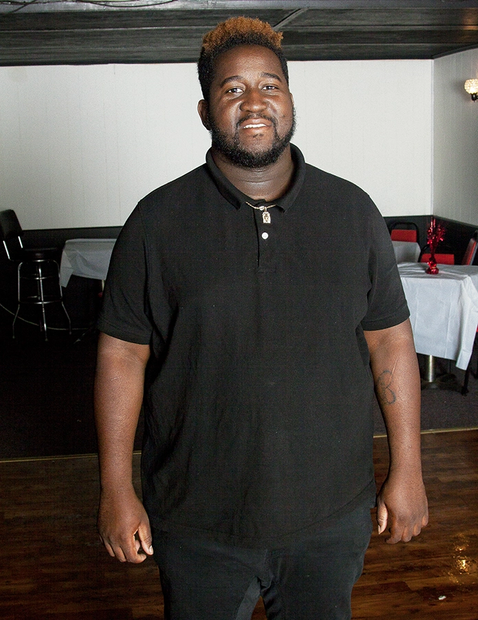 Smiling man in a black polo shirt stands in a dimly lit venue with tables and chairs in the background.